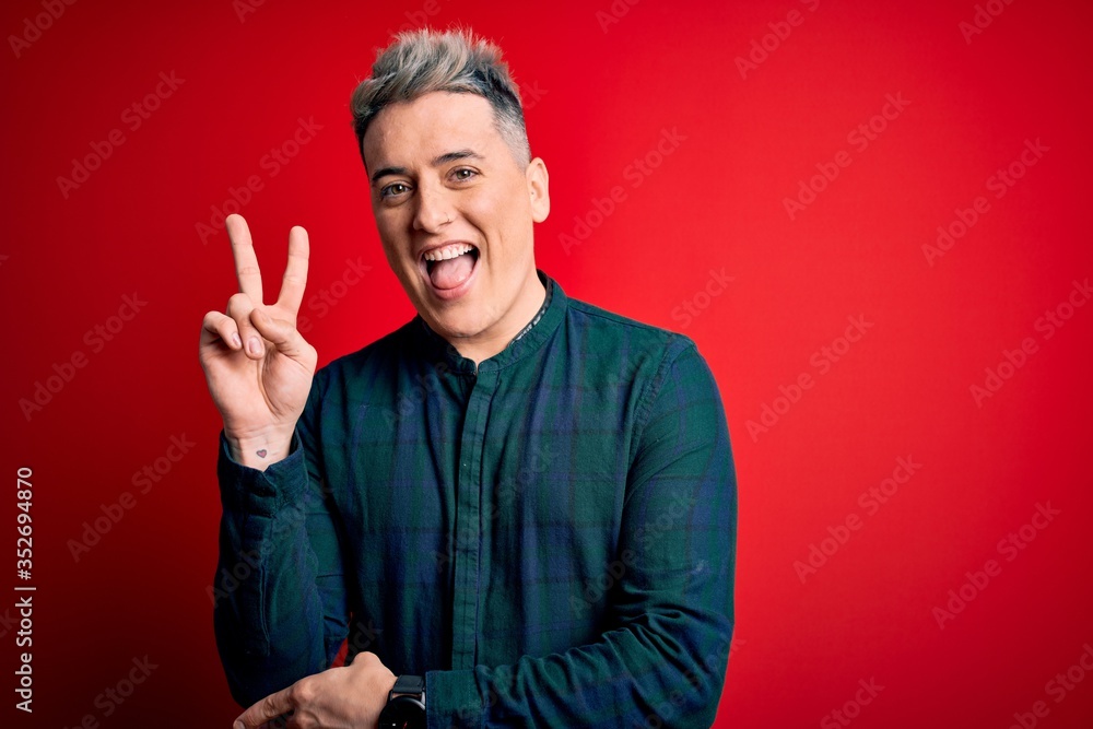 Young handsome modern man wearing elegant green shirt over red isolated background smiling with happy face winking at the camera doing victory sign with fingers. Number two. Young handsome modern man wearing elegant green shirt over red isolated background smiling with happy face winking at the camera doing victory sign with fingers. Number two.