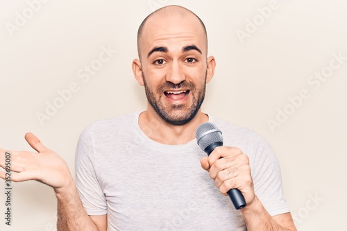 Young handsome bald man singing song using microphone celebrating achievement with happy smile and winner expression with raised hand