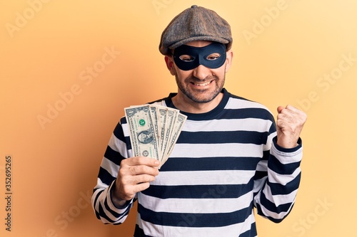 Young handsome bald man wearing burglar mask stealing dollars banknotes screaming proud, celebrating victory and success very excited with raised arm photo