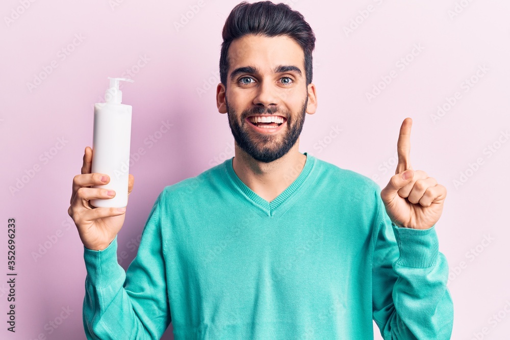 Young handsome man with beard holding bottle of cream smiling with an idea or question pointing finger with happy face, number one Young handsome man with beard holding bottle of cream smiling with an idea or question pointing finger with happy face, number one