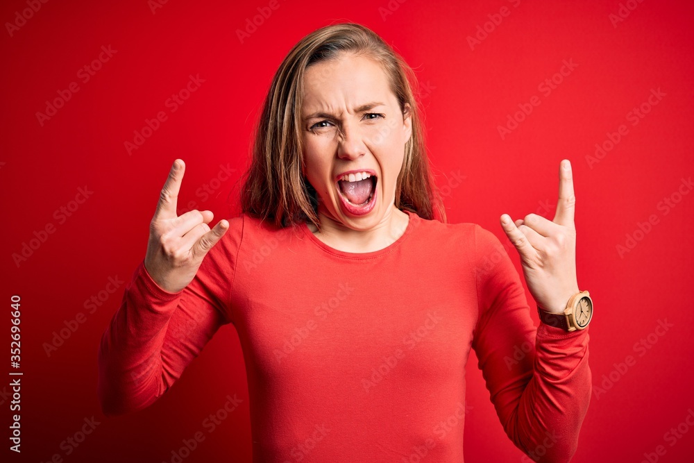 Young beautiful blonde woman wearing casual t-shirt standing over isolated red background shouting with crazy expression doing rock symbol with hands up. Music star. Heavy concept. Young beautiful blonde woman wearing casual t-shirt standing over isolated red background shouting with crazy expression doing rock symbol with hands up. Music star. Heavy concept.