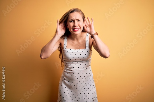 Young beautiful blonde woman on vacation wearing summer dress over yellow background Trying to hear both hands on ear gesture, curious for gossip. Hearing problem, deaf