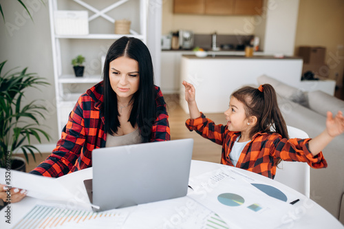 Distant work. Young mom try to work remotely on laptop at home, and her cute cheerful daughter interferes with her and distracts photo