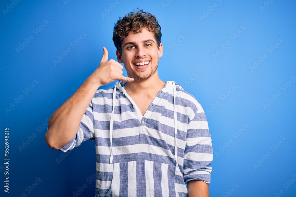 Young blond handsome man with curly hair wearing casual striped sweatshirt smiling doing phone gesture with hand and fingers like talking on the telephone. Communicating concepts. Young blond handsome man with curly hair wearing casual striped sweatshirt smiling doing phone gesture with hand and fingers like talking on the telephone. Communicating concepts.