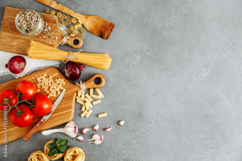 Spaghetti and fettuccine with ingredients for cooking pasta on cement backdrop with blank of wooden kitchen board, top view. Rustic style. Flat lay with space for text