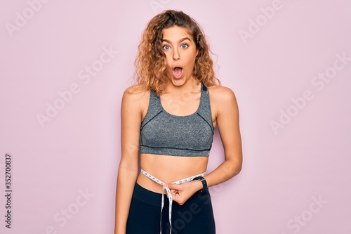 Young blonde sporty woman with blue eyes controlling weight using tape measure on waist scared in shock with a surprise face, afraid and excited with fear expression © Krakenimages.com
