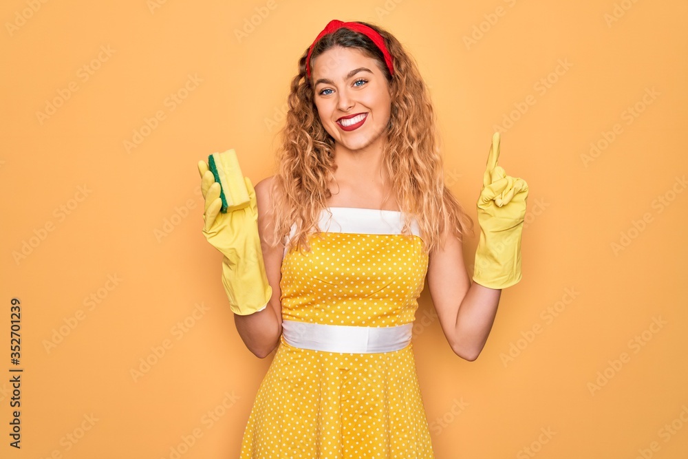 Young beautiful blonde woman with blue eyes cleaning using gloves and cleaner scourer surprised with an idea or question pointing finger with happy face, number one Young beautiful blonde woman with blue eyes cleaning using gloves and cleaner scourer surprised with an idea or question pointing finger with happy face, number one