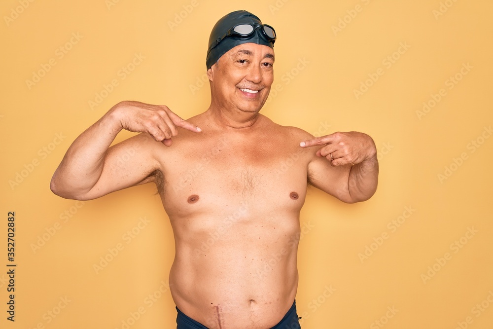 Middle age senior grey-haired swimmer man wearing swimsuit, cap and goggles looking confident with smile on face, pointing oneself with fingers proud and happy.