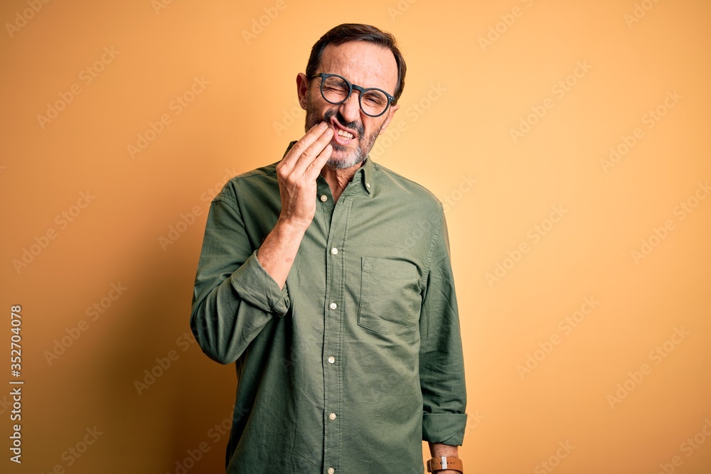 Middle age hoary man wearing casual green shirt and glasses over isolated yellow background touching mouth with hand with painful expression because of toothache or dental illness on teeth. Dentist Middle age hoary man wearing casual green shirt and glasses over isolated yellow background touching mouth with hand with painful expression because of toothache or dental illness on teeth. Dentist