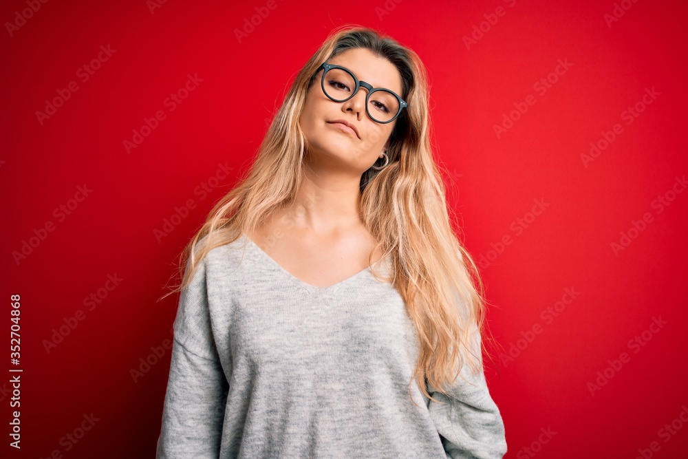 Young beautiful blonde woman wearing sweater and glasses over isolated red background Relaxed with serious expression on face. Simple and natural looking at the camera.