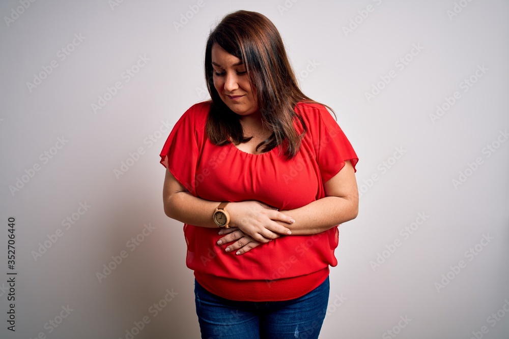 Beautiful brunette plus size woman wearing casual red t-shirt over isolated white background with hand on stomach because indigestion, painful illness feeling unwell. Ache concept. Beautiful brunette plus size woman wearing casual red t-shirt over isolated white background with hand on stomach because indigestion, painful illness feeling unwell. Ache concept.