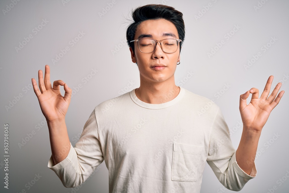 Young handsome chinese man wearing casual t-shirt and glasses over white background relax and smiling with eyes closed doing meditation gesture with fingers. Yoga concept. Young handsome chinese man wearing casual t-shirt and glasses over white background relax and smiling with eyes closed doing meditation gesture with fingers. Yoga concept.