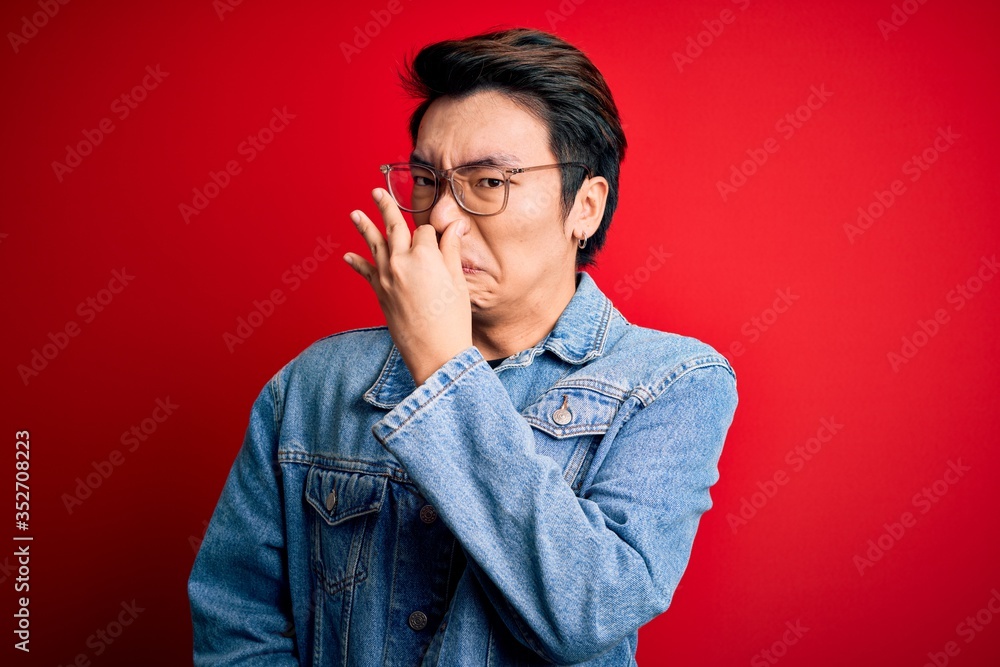 Young handsome chinese man wearing denim jacket and glasses over red background smelling something stinky and disgusting, intolerable smell, holding breath with fingers on nose. Bad smell Young handsome chinese man wearing denim jacket and glasses over red background smelling something stinky and disgusting, intolerable smell, holding breath with fingers on nose. Bad smell