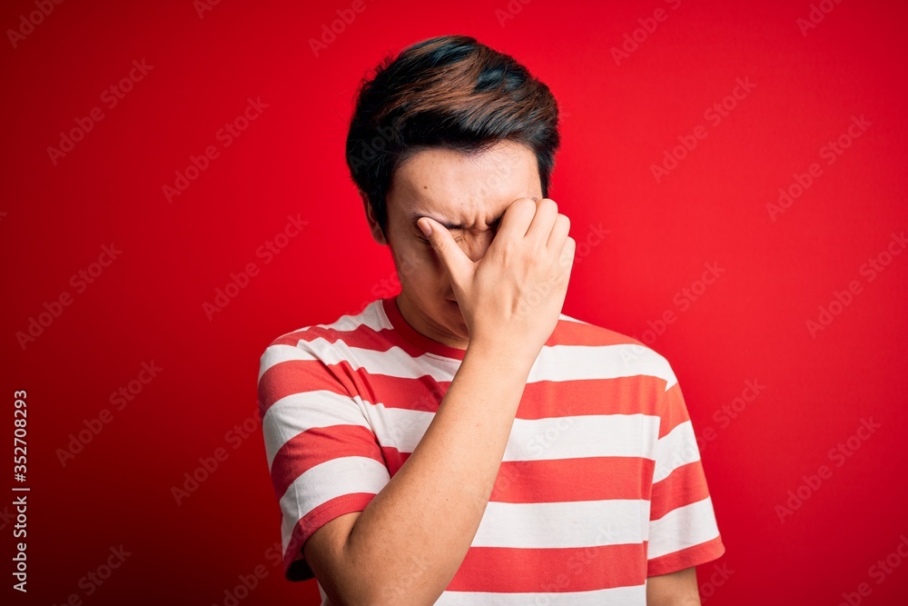 Young handsome chinese man wearing casual striped t-shirt standing over red background tired rubbing nose and eyes feeling fatigue and headache. Stress and frustration concept. Young handsome chinese man wearing casual striped t-shirt standing over red background tired rubbing nose and eyes feeling fatigue and headache. Stress and frustration concept.