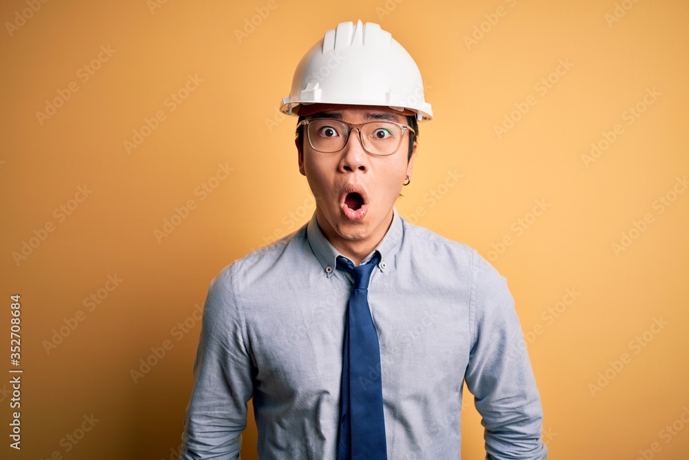 Young handsome chinese architect man wearing safety helmet and tie over yellow background afraid and shocked with surprise expression, fear and excited face. Young handsome chinese architect man wearing safety helmet and tie over yellow background afraid and shocked with surprise expression, fear and excited face.