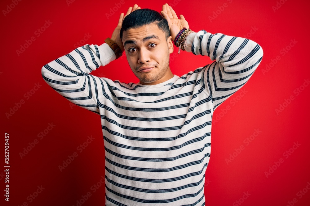 Young brazilian man wearing casual striped t-shirt standing over isolated red background Doing bunny ears gesture with hands palms looking cynical and skeptical. Easter rabbit concept. Young brazilian man wearing casual striped t-shirt standing over isolated red background Doing bunny ears gesture with hands palms looking cynical and skeptical. Easter rabbit concept.