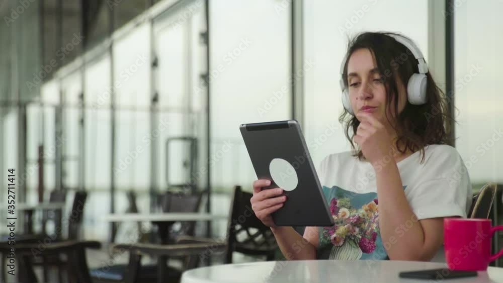 Thoughtful woman with headphones is sitting at cafe using tablet Thoughtful woman with headphones is sitting at cafe using tablet
