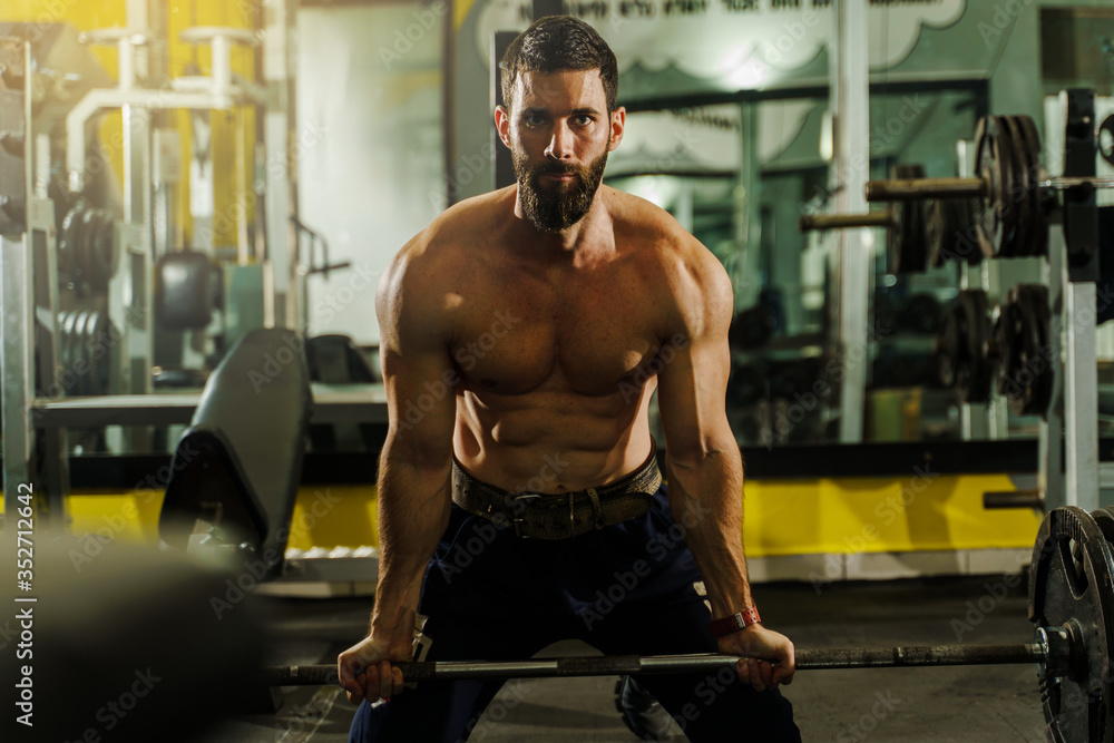 Front view portrait of adult caucasian man male athlete bodybuilder muscular handsome shirtless holding barbel with weights in dark gym while training weight lifting deadlift bodybuilding or fitness Front view portrait of adult caucasian man male athlete bodybuilder muscular handsome shirtless holding barbel with weights in dark gym while training weight lifting deadlift bodybuilding or fitness