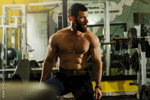 Front view portrait of adult caucasian man male athlete muscular bodybuilder or fitness trainer with black hair and beard sitting shirtless at the gym looking to the side resting