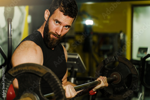 Side view portrait of young caucasian man male athlete bodybuilder training at the gym workout using barbel and weights weight lifting biceps curls wearing black shirt dark hair and beard standing