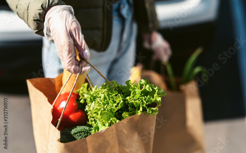 A young woman with grocery bags from a supermarket. Car trunk at background. Social distancing: face mask, disposable gloves to prevent infection. Food shopping during coronavirus Covid 19 epidemic