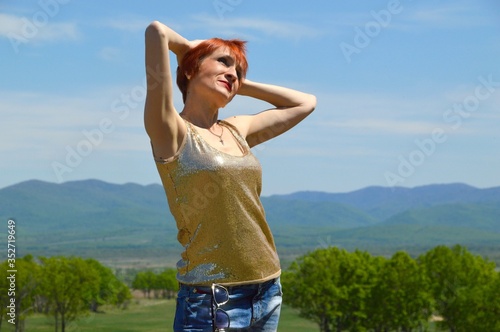 Red-haired cheerful woman smilling on the background of a green field on a summer sunny day. She touching her hair with hands and looking away. Enjoying freedom