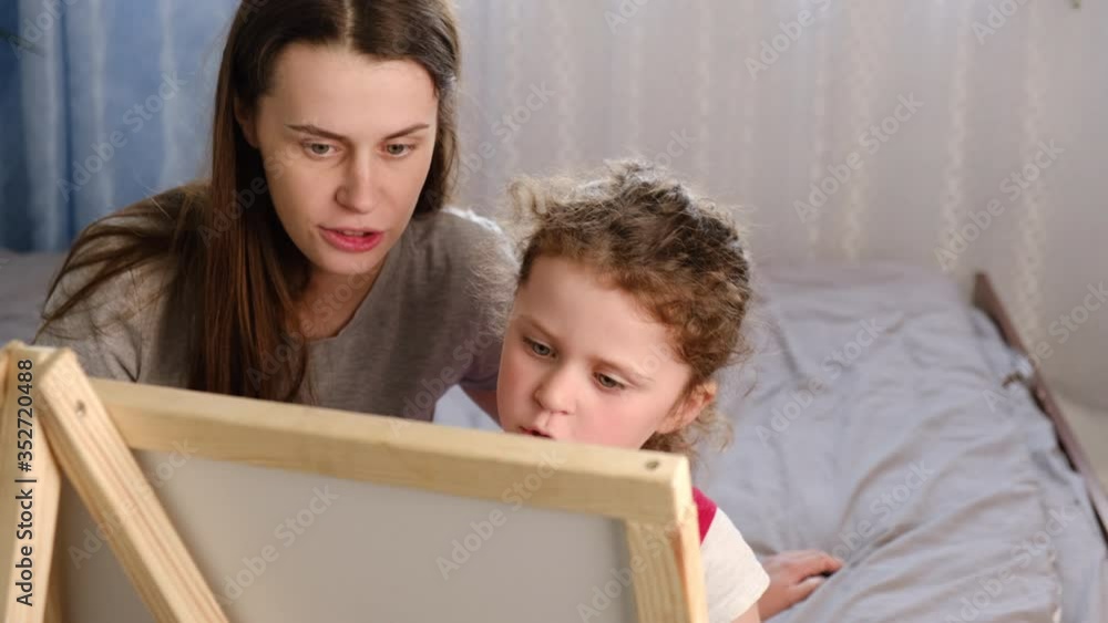 Young mother teaches preschool daughter to write words with chalk on a small blackboard sitting on bed at home. Little girl learning writing with mom tutor. Children education concept Young mother teaches preschool daughter to write words with chalk on a small blackboard sitting on bed at home. Little girl learning writing with mom tutor. Children education concept