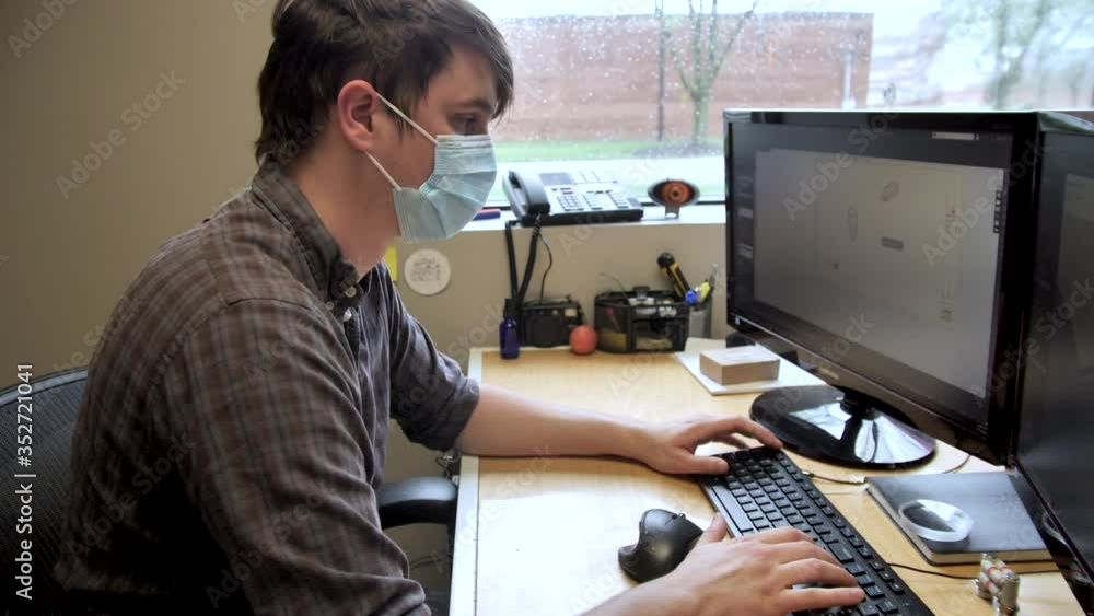 Young Caucasian man works on office computer at work wearing face mask, covid-19 pandemic, close up Young Caucasian man works on office computer at work wearing face mask, covid-19 pandemic, close up