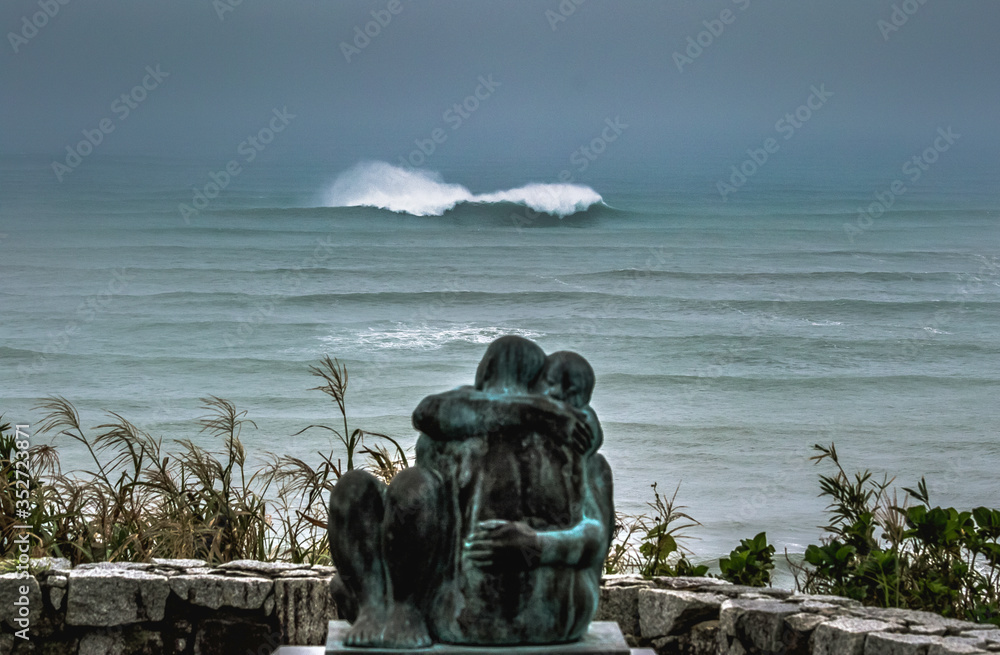 Typhoon in Japan, a couple hugging statue with a large wave breaking in the background out at sea, with a stormy sky, taken in Katsuura, Chiba, Japan near to Hebara Beach which is famous for surfing a
