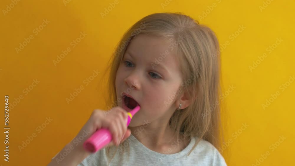 Charming little girl in grey t-shirt cleaning teeth with colorful kids toothbrush looking at camera and dancing on yellow background Charming little girl in grey t-shirt cleaning teeth with colorful kids toothbrush looking at camera and dancing on yellow background