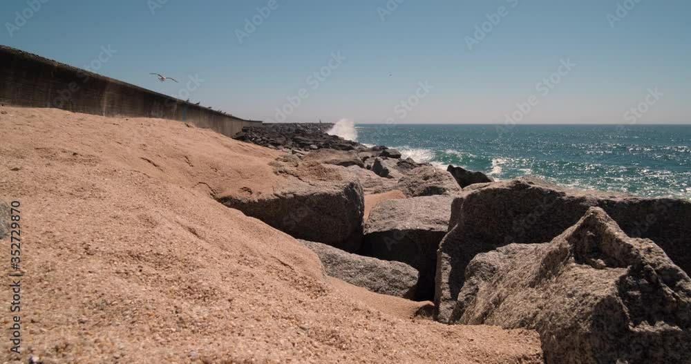 Waves breaking against sea wall in Povoa de Varzim, Portugal as seagulls fly overhead. Bright sunny summer day with blue sky. Waves breaking against sea wall in Povoa de Varzim, Portugal as seagulls fly overhead. Bright sunny summer day with blue sky.