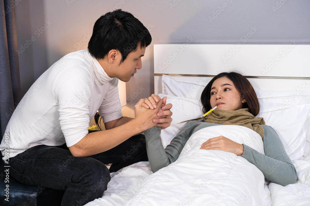 Obraz premium sick woman suffering in bed at home, her husband checking temperature with thermometer.