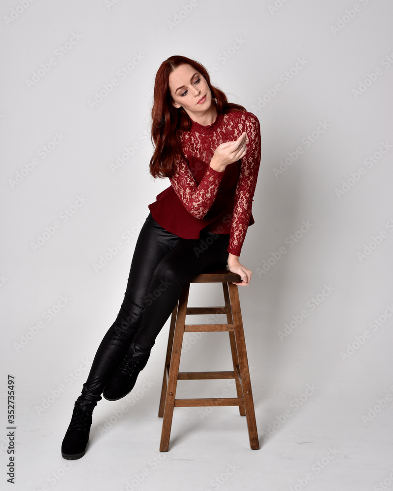 Portrait of a pretty girl with red hair wearing leather pants and long sleeved lace shirt. full length sitting pose on wooden stool, isolated against a studio background Portrait of a pretty girl with red hair wearing leather pants and long sleeved lace shirt. full length sitting pose on wooden stool, isolated against a studio background