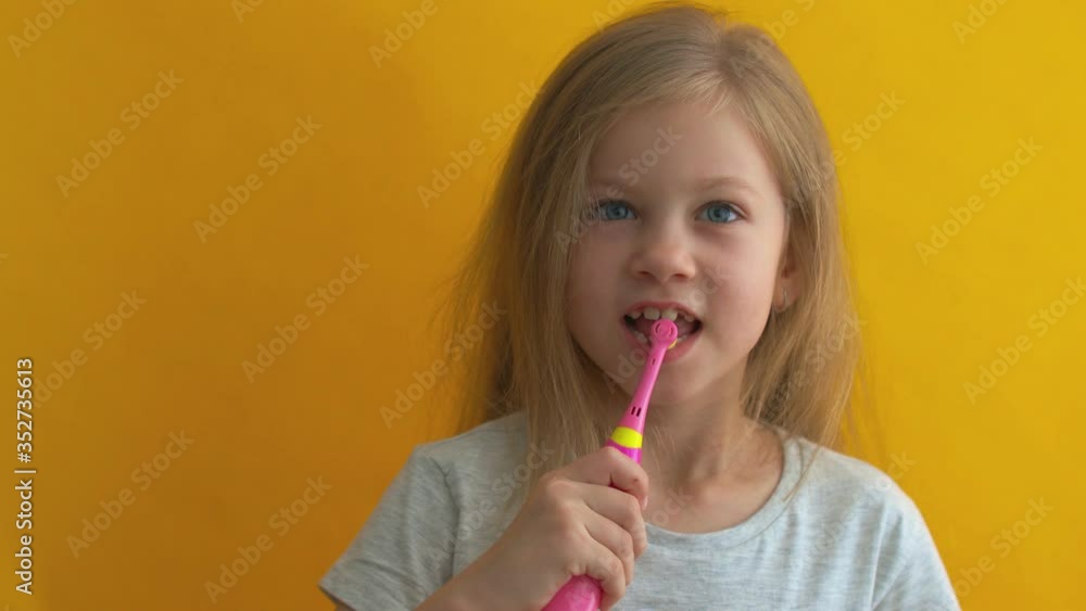 Charming little girl in grey t-shirt cleaning teeth with electric kids toothbrush looking at camera on a yellow background. Charming little girl in grey t-shirt cleaning teeth with electric kids toothbrush looking at camera on a yellow background.