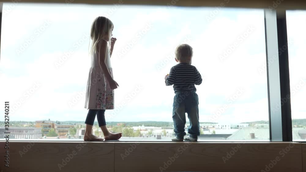 Young sister and brother standing in big hotel window looking down on the city Young sister and brother standing in big hotel window looking down on the city