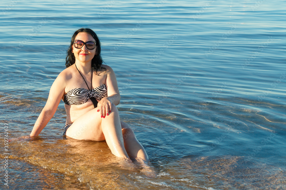 A brunette woman in a swimsuit and glasses sits on the seashore.