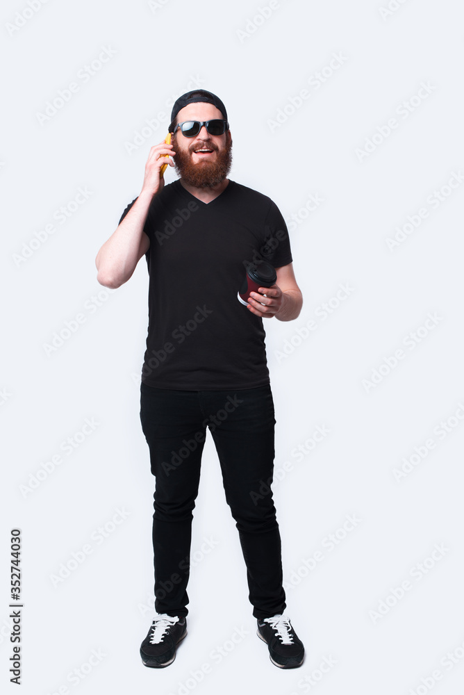 Full length photo of man with beard talking on phone and holding coffee Full length photo of man with beard talking on phone and holding coffee
