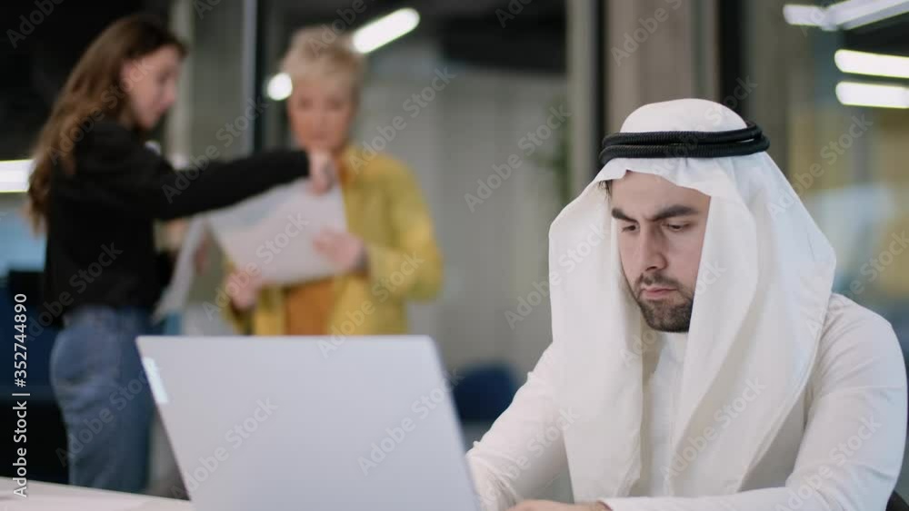 Arab businessman is working on a laptop intently, next to him are two businesswomen discussing documentation. Arab businessman is working on a laptop intently, next to him are two businesswomen discussing documentation.
