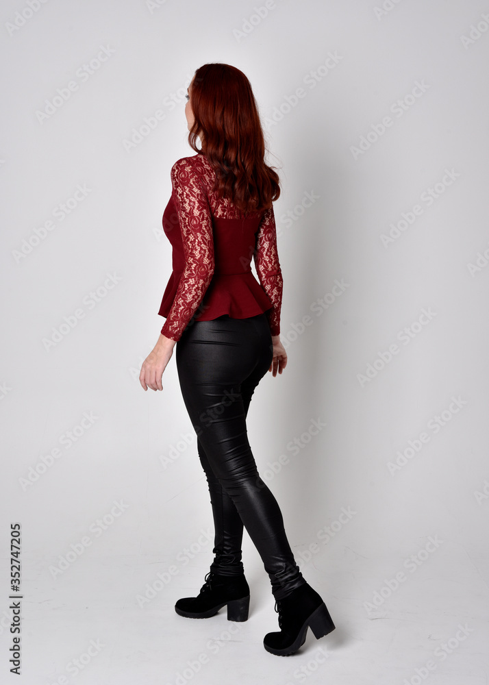 Portrait of a pretty girl with red hair wearing leather pants and long sleeved lace shirt. full length standing pose facing away from the camera, isolated against a studio background. Portrait of a pretty girl with red hair wearing leather pants and long sleeved lace shirt. full length standing pose facing away from the camera, isolated against a studio background.