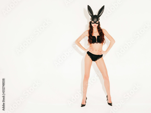 Beautiful sexy woman wearing carnival black mask of Easter bunny rabbit.Hot blonde girl posing near white wall in studio. Seductive model in nice lingerie