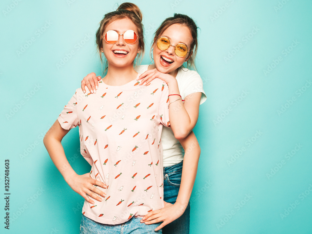 Two young beautiful blond smiling hipster girls in trendy summer hipster jeans clothes. Sexy carefree women posing near blue wall. Trendy and positive models having fun in sunglasses Two young beautiful blond smiling hipster girls in trendy summer hipster jeans clothes. Sexy carefree women posing near blue wall. Trendy and positive models having fun in sunglasses