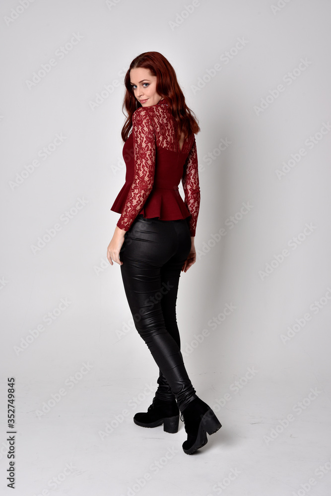 Portrait of a pretty girl with red hair wearing leather pants and long sleeved lace shirt. full length standing pose facing away from the camera, isolated against a studio background. Portrait of a pretty girl with red hair wearing leather pants and long sleeved lace shirt. full length standing pose facing away from the camera, isolated against a studio background.
