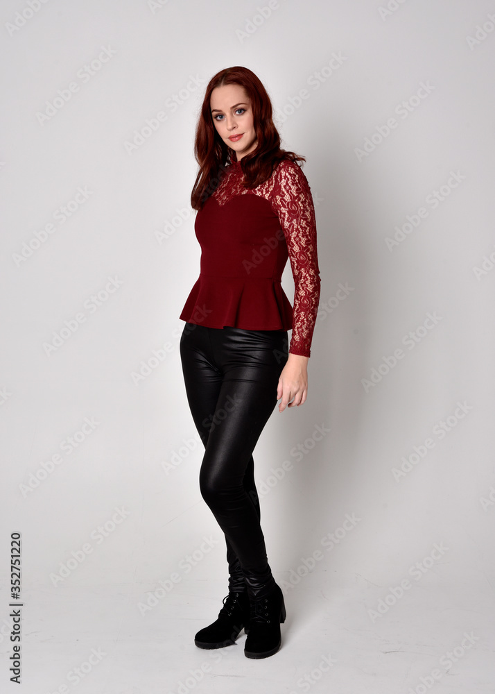 Portrait of a pretty girl with red hair wearing leather pants and long sleeved lace shirt. full length standing pose, isolated against a studio background. Portrait of a pretty girl with red hair wearing leather pants and long sleeved lace shirt. full length standing pose, isolated against a studio background.