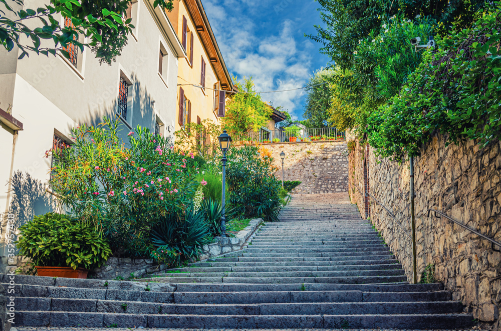 Cobblestone staircase with stairs, green trees, bushes and flowers, street lights between stone walls in Brescia city historical centre, Lombardy, Northern Italy