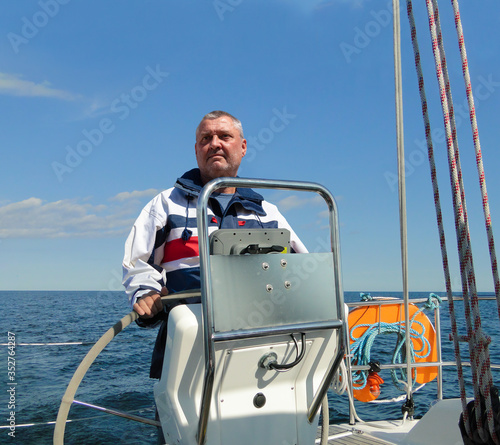 The captain at the wheel of a sailing yacht in calm seas, sunshine and blue skies. The man with the beard looks at the sea. He wears waterproof sailing clothing. Behind the man is a lifebuoy.