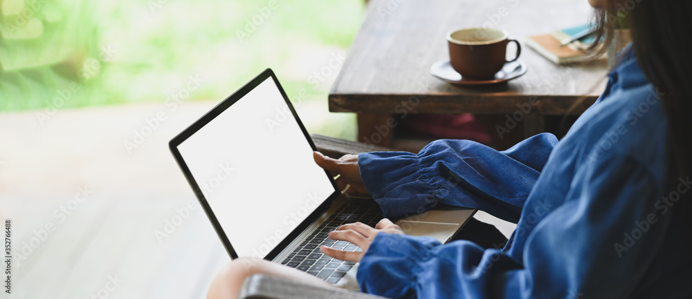 Photo of beautiful woman typing on white blank screen computer laptop that putting on her lap and sitting next to wooden desk outside the house over nature as background. - obrazy, fototapety, plakaty 