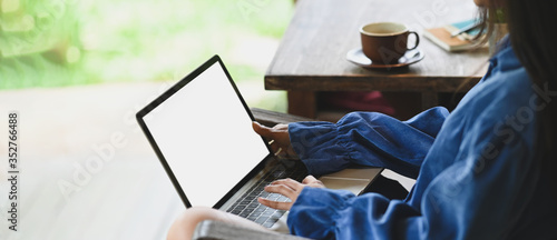 Photo of beautiful woman typing on white blank screen computer laptop that putting on her lap and sitting next to wooden desk outside the house over nature as background.