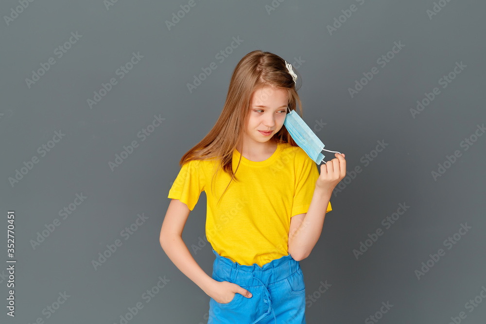 A little girl stands with a mask in her hand on a gray background, annoyed by the mask, she wants to take it off A little girl stands with a mask in her hand on a gray background, annoyed by the mask, she wants to take it off