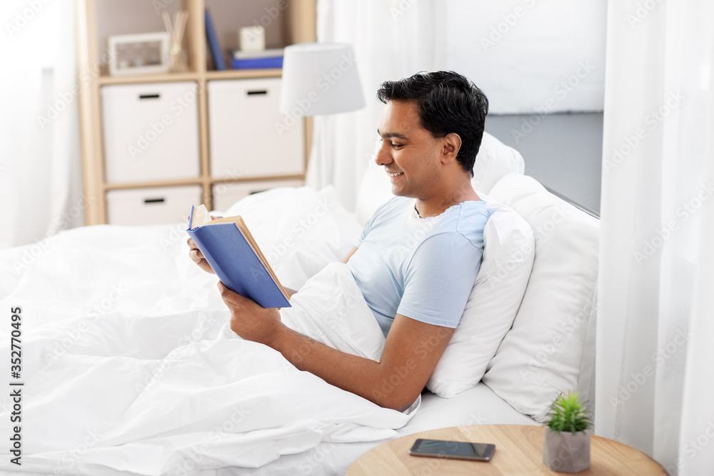 Fototapeta premium people, bedtime and rest concept - happy smiling indian man reading book in bed at home