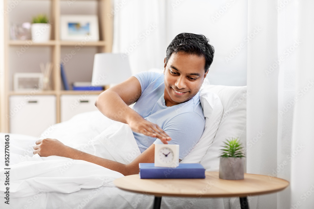 people, bedtime and rest concept - happy smiling indian man with alarm clock lying in bed at home people, bedtime and rest concept - happy smiling indian man with alarm clock lying in bed at home